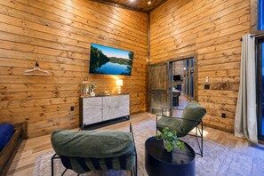A cozy room with wooden walls, a large TV mounted on the wall, a cabinet with decorative items, and two chairs with a small table in the center. - Broken Bow Bliss · Private Heated Pool, Hot Tub & Arcade Game Room | Sleeps 12 (Broken Bow)