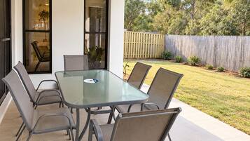 Une terrasse extérieure avec une table à plateau de verre et six chaises, une clôture en bois en arrière-plan et une vue sur les arbres.