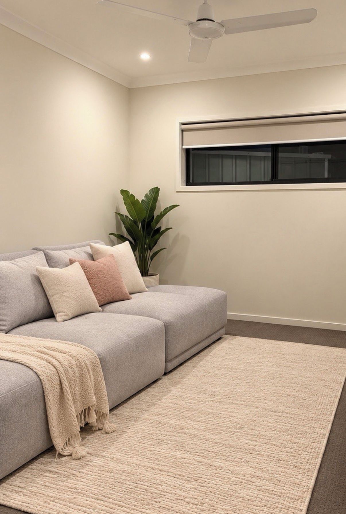 Un salon moderne avec un canapé gris, un tapis beige, un ventilateur de plafond et une fenêtre équipée de stores.