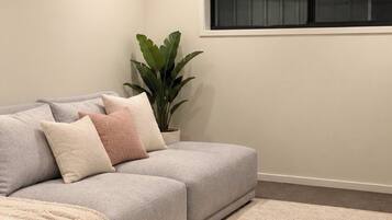 Un salon moderne avec un canapé gris, un tapis beige, un ventilateur de plafond et une fenêtre équipée de stores.