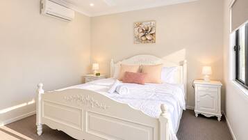 Une chambre à coucher avec un lit blanc, deux tables de chevet avec des lampes, un ventilateur de plafond et un climatiseur fixé au mur.