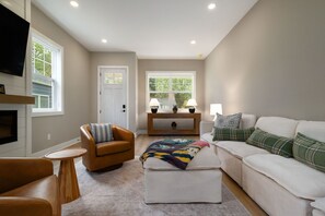 A living room with a brown leather chair, a white sofa, a wooden coffee table, and a television mounted on the wall. - Irish Victory House | 5BR/4BA (South Bend)