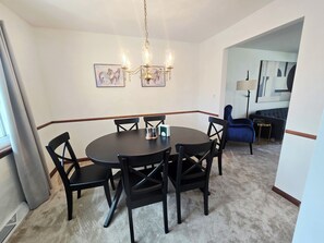 Un coin repas avec une table ronde, six chaises et un lustre. Deux tableaux encadrés ornent le mur.