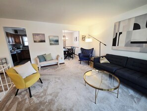 Un salon comprenant un canapé, un fauteuil et une table basse, un coin repas avec des chaises, et une cuisine visible à travers une porte ouverte.