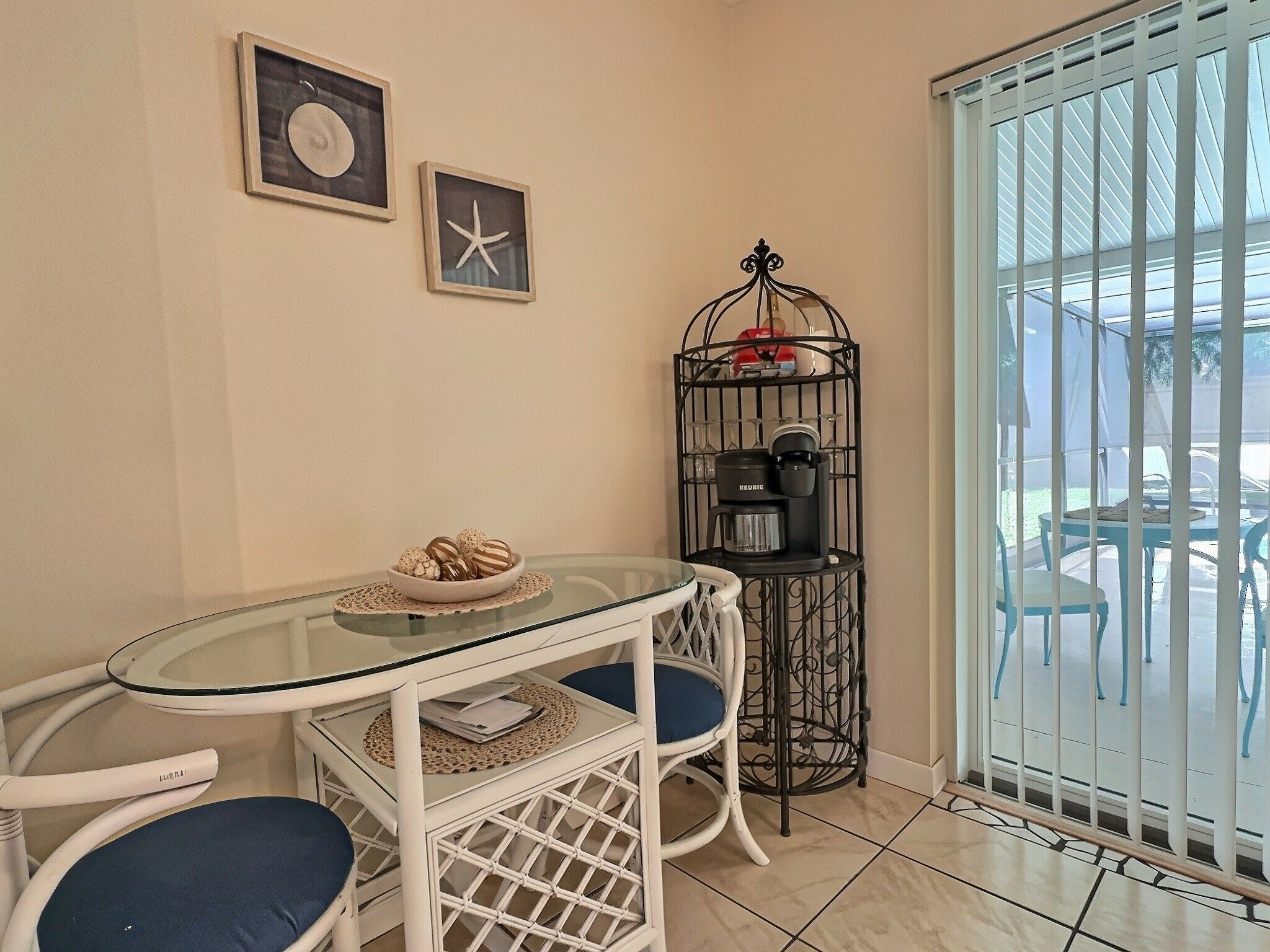 A corner room with a glass table, two chairs, a coffee machine, and a view through a window with a metal gate.