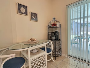 Habitación de esquina con mesa de vidrio, dos sillas, una cafetera y vista a través de una ventana con reja.