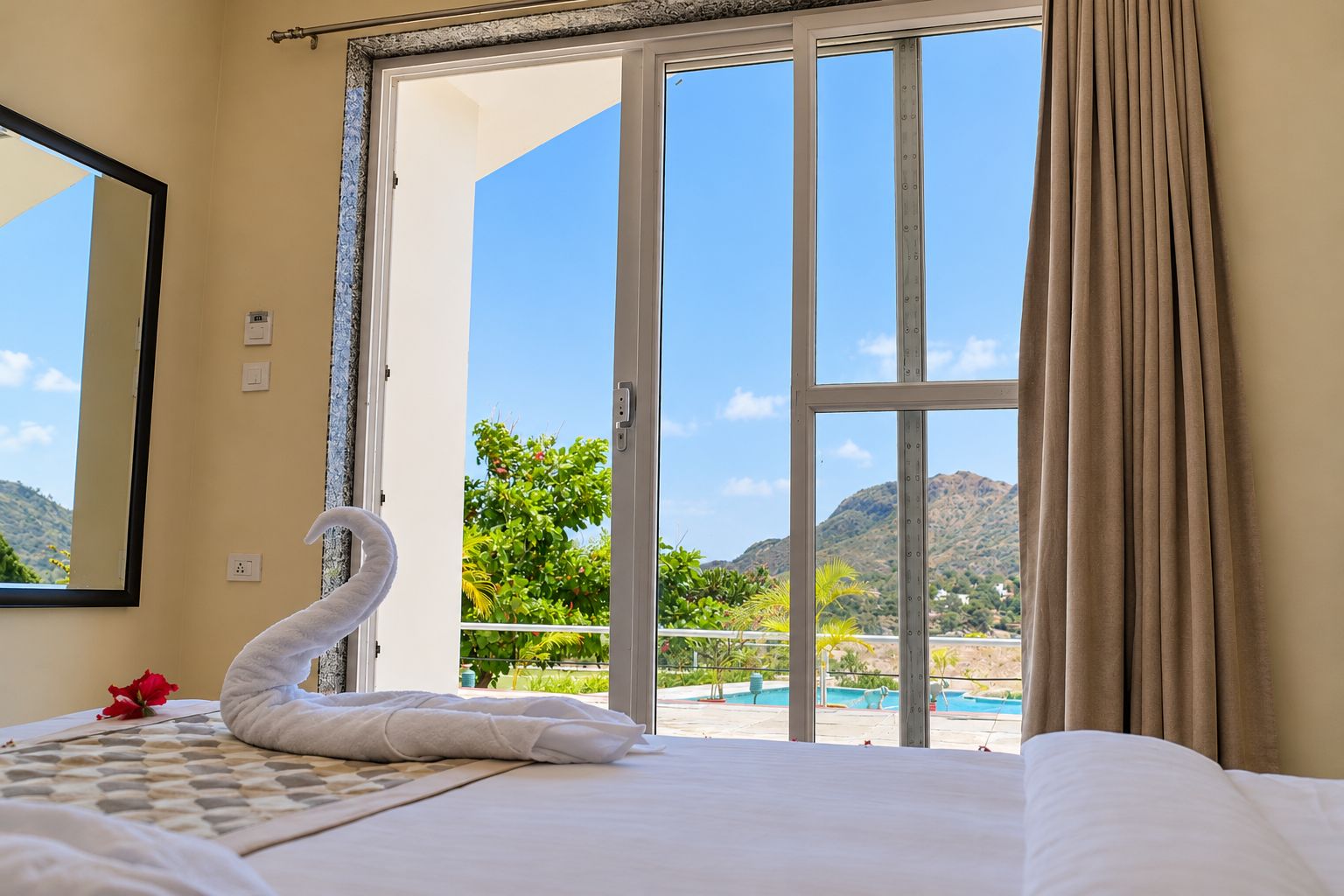 A hotel room with a bed, a swan-shaped towel, a view of a pool and mountains.