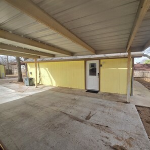 Un espace extérieur abrité, doté d’un sol en béton, d’un mur jaune et d’une porte blanche.