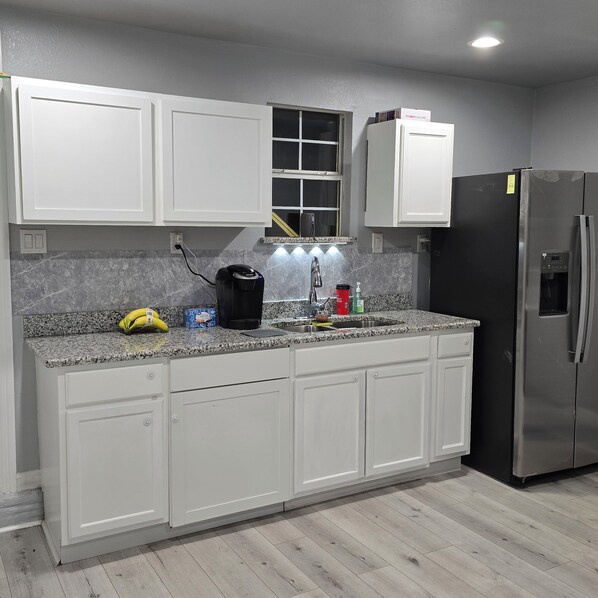 A kitchen with white cabinets, a granite countertop, a black coffee maker, a red cup, and a stainless steel refrigerator.