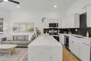 A modern kitchen with white cabinetry, a central island, and a dining area with a sofa and table. - Large Group Stay | Walk to St Augustine Downtown (St. Augustine)