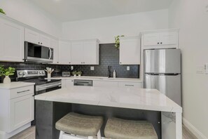 A modern kitchen with white cabinetry, a stainless steel refrigerator, and a central island with stools. - Large Group Stay | Walk to St Augustine Downtown (St. Augustine)