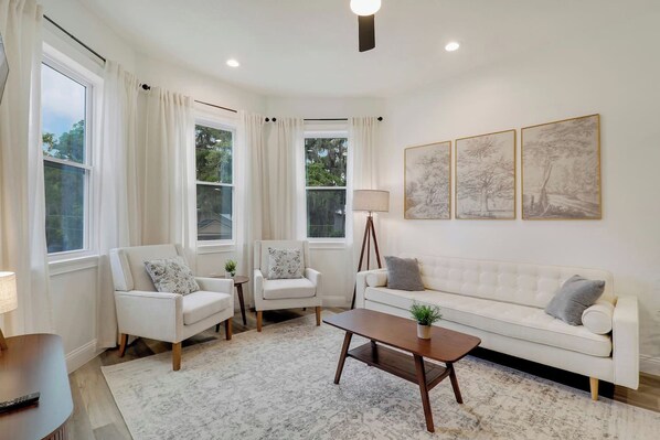 A modern living room with a white sofa, two armchairs, a coffee table, and a floor lamp. There are three large windows with curtains, and three framed artworks on the wall. - Walk to St Augustine | Fully Renovated Luxury Home (St. Augustine)