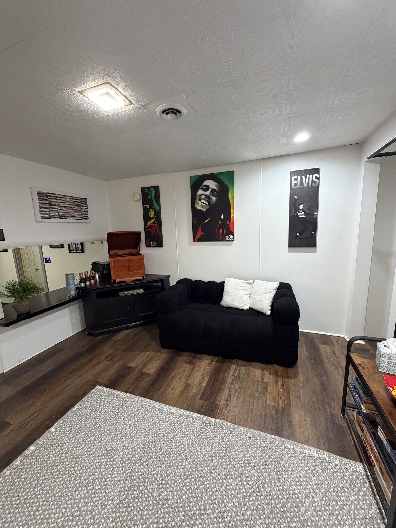 A modern living room with a black sofa, a coffee table, and a wooden cabinet. There are posters on the wall and a small plant on the counter.
