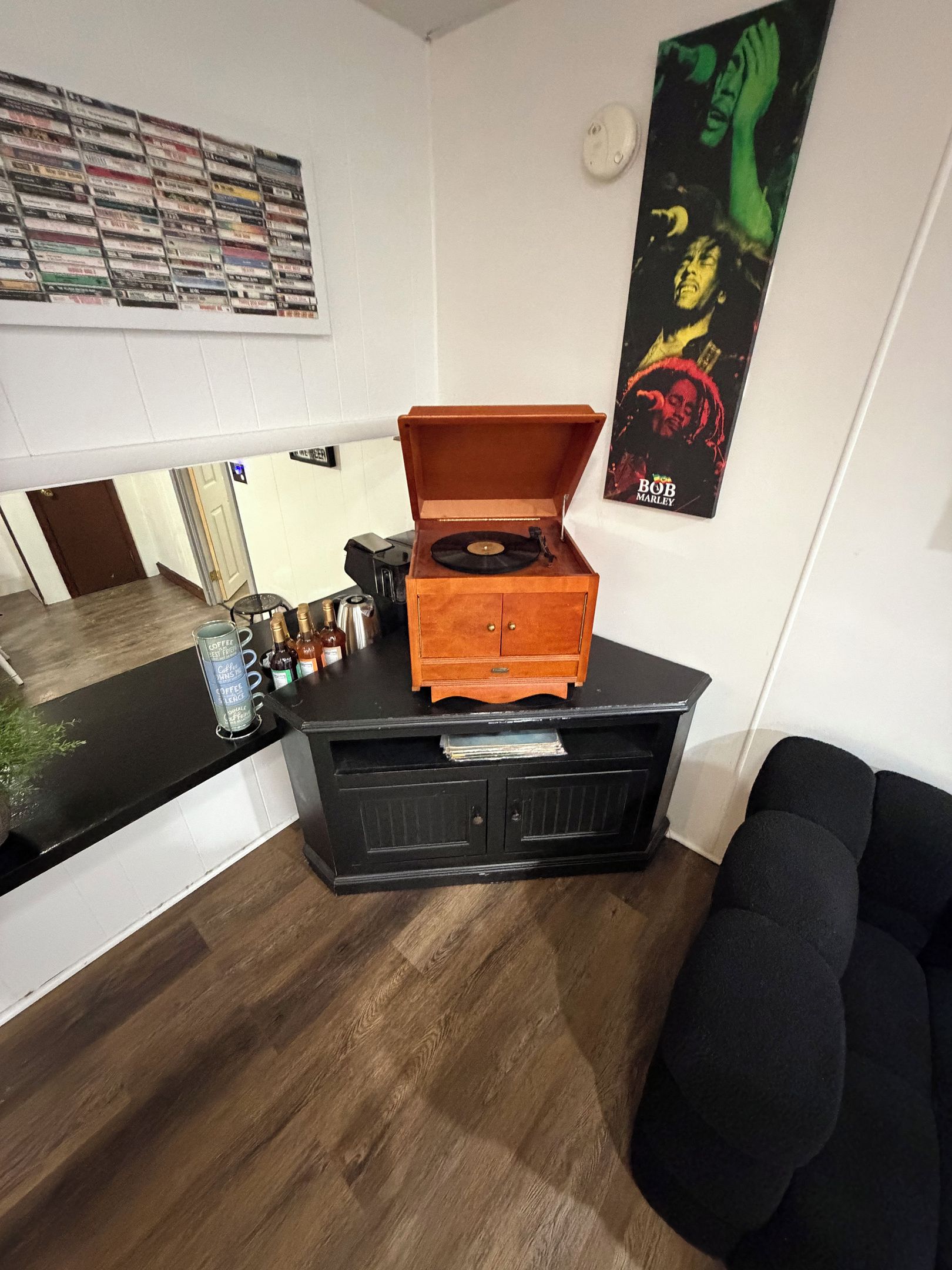 A room with a wooden cabinet containing a record player, a coffee machine, and various bottles. There is a poster on the wall and a black couch in the corner.