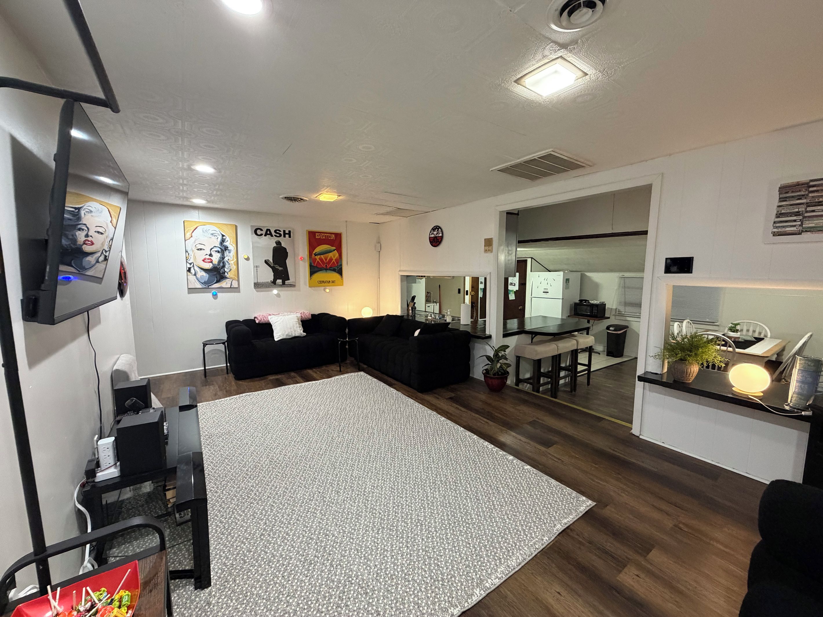 Ein modernes Wohnzimmer mit großem Teppich, schwarzen Sofas, einem Flachbildfernseher und einem durch einen offenen Durchgang einsehbarer Küchenbereich.