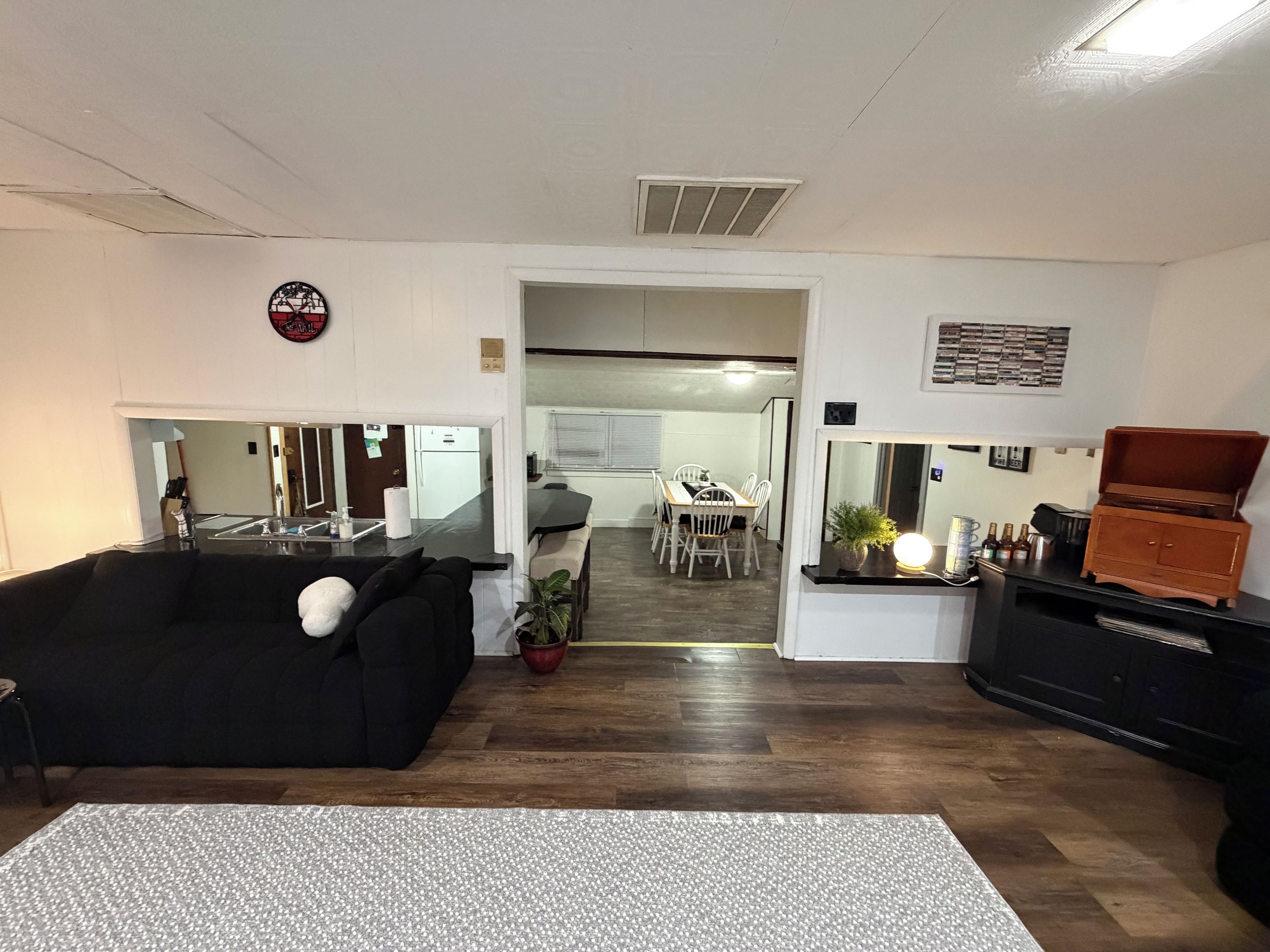 Una sala de estar con un sofá negro, una mesa de comedor con sillas, una zona de cocina y un mueble de madera pequeño.