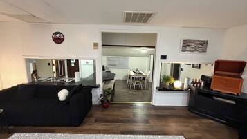 Una sala de estar con un sofá negro, una mesa de comedor con sillas, una zona de cocina y un mueble de madera pequeño.