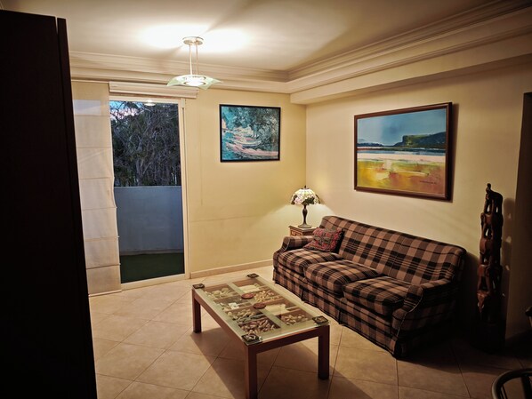 A living room with a sofa, a coffee table, and a lamp. There are two paintings on the wall and a window with a view of trees.