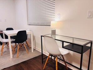 Ein Zimmer mit weißem Schreibtisch, zwei Stühlen, einem Glastisch, einer Lampe und einem Fenster mit Jalousien.