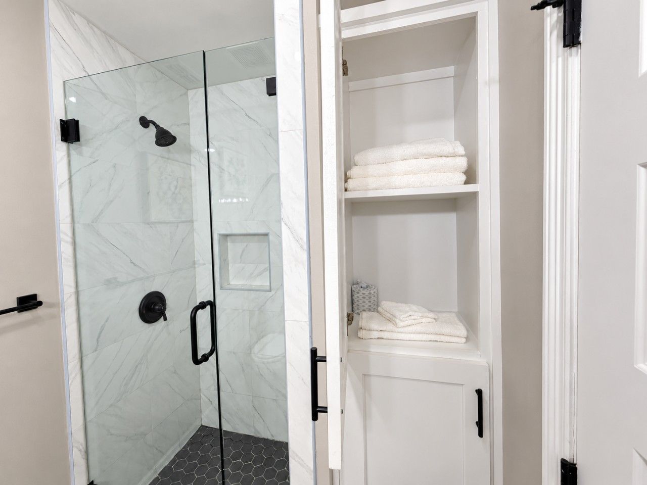 A bathroom with a glass shower enclosure, a white cabinet with towels, and a door with a black handle.