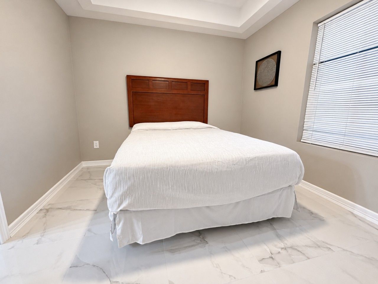 Une chambre à coucher avec un lit, une tête de lit en bois, un sol en marbre, une fenêtre avec des stores et un tableau encadré au mur.
