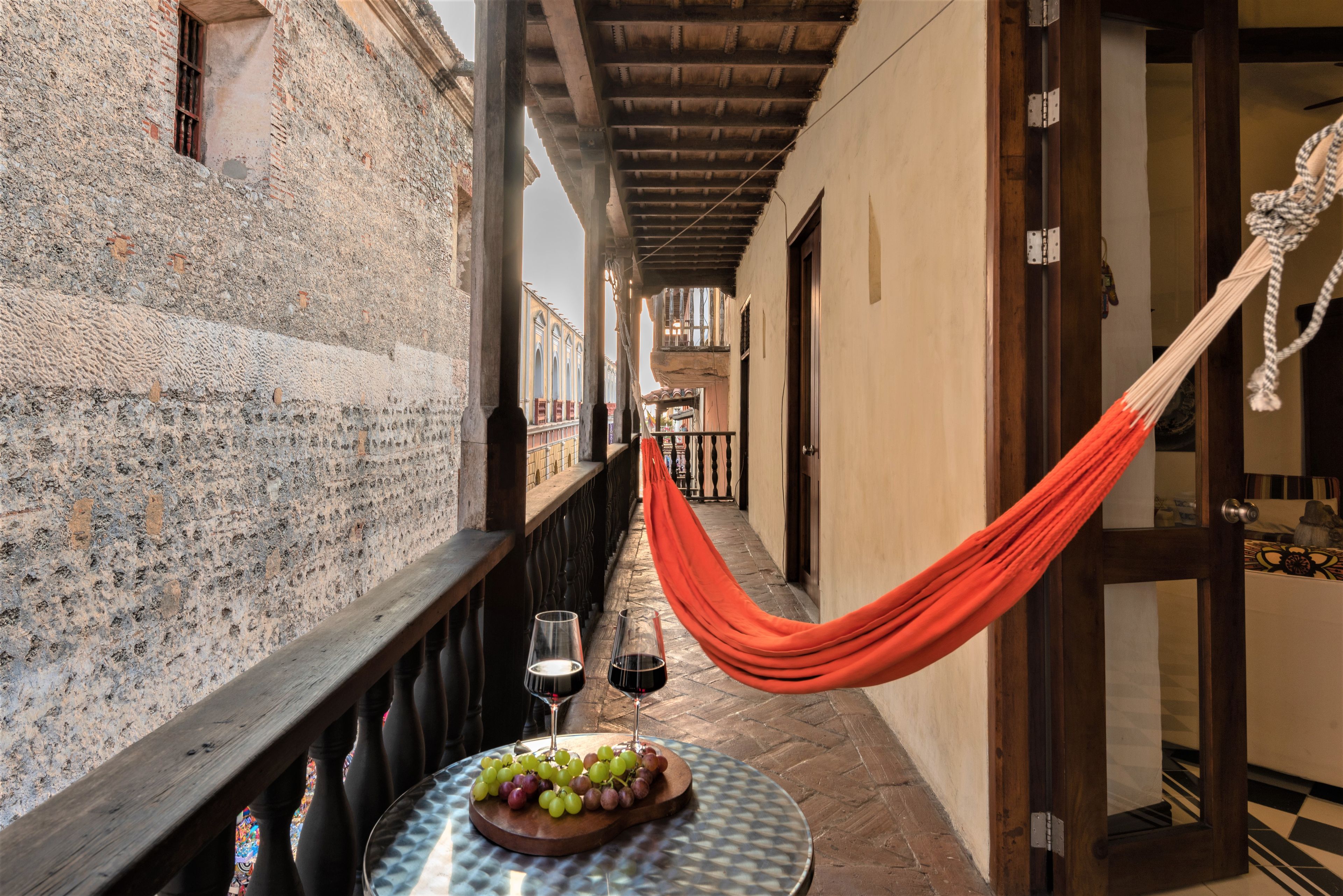 Un couloir étroit avec un hamac, une table sur laquelle est disposée une assiette de fruits et des verres à vin, et une vue sur un bâtiment à la façade recouverte de carreaux.