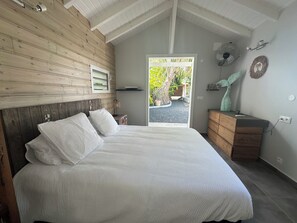 A bedroom with a wooden headboard, a bed with white bedding, a nightstand with a lamp, a dresser, and a fan on the wall. - New Listing (Saint-François)