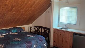 A bedroom with a sloped ceiling, a bed with a floral bedspread, a wooden chair, a small kitchenette with a sink, and a window with blinds.