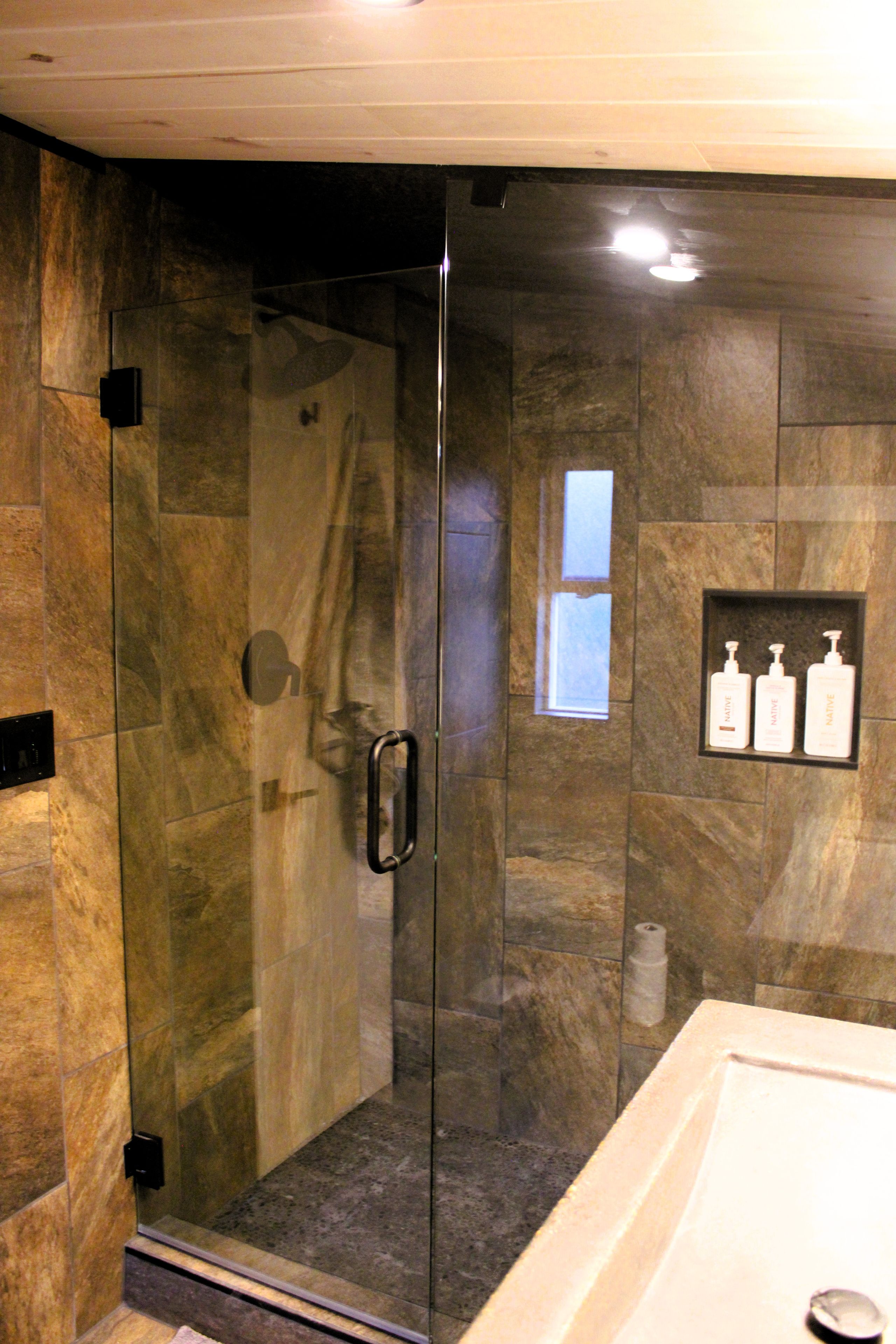 A glass-enclosed shower with a handrail, a marble-tiled wall, and a shelf with toiletry bottles.