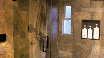 A glass-enclosed shower with a handrail, a marble-tiled wall, and a shelf with toiletry bottles.