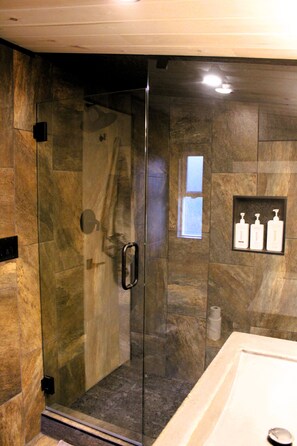 A glass-enclosed shower with a handrail, a marble-tiled wall, and a shelf with toiletry bottles.