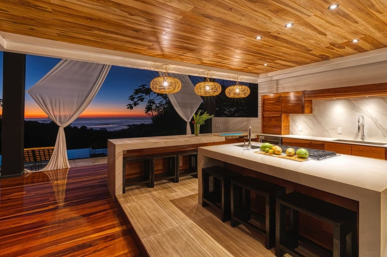 A modern kitchen with a wooden ceiling, large windows, and a view of the sunset.