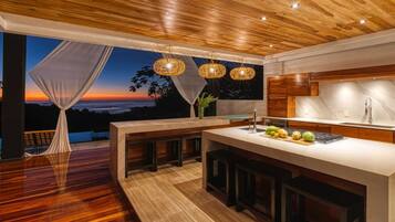 A modern kitchen with a wooden ceiling, large windows, and a view of the sunset.