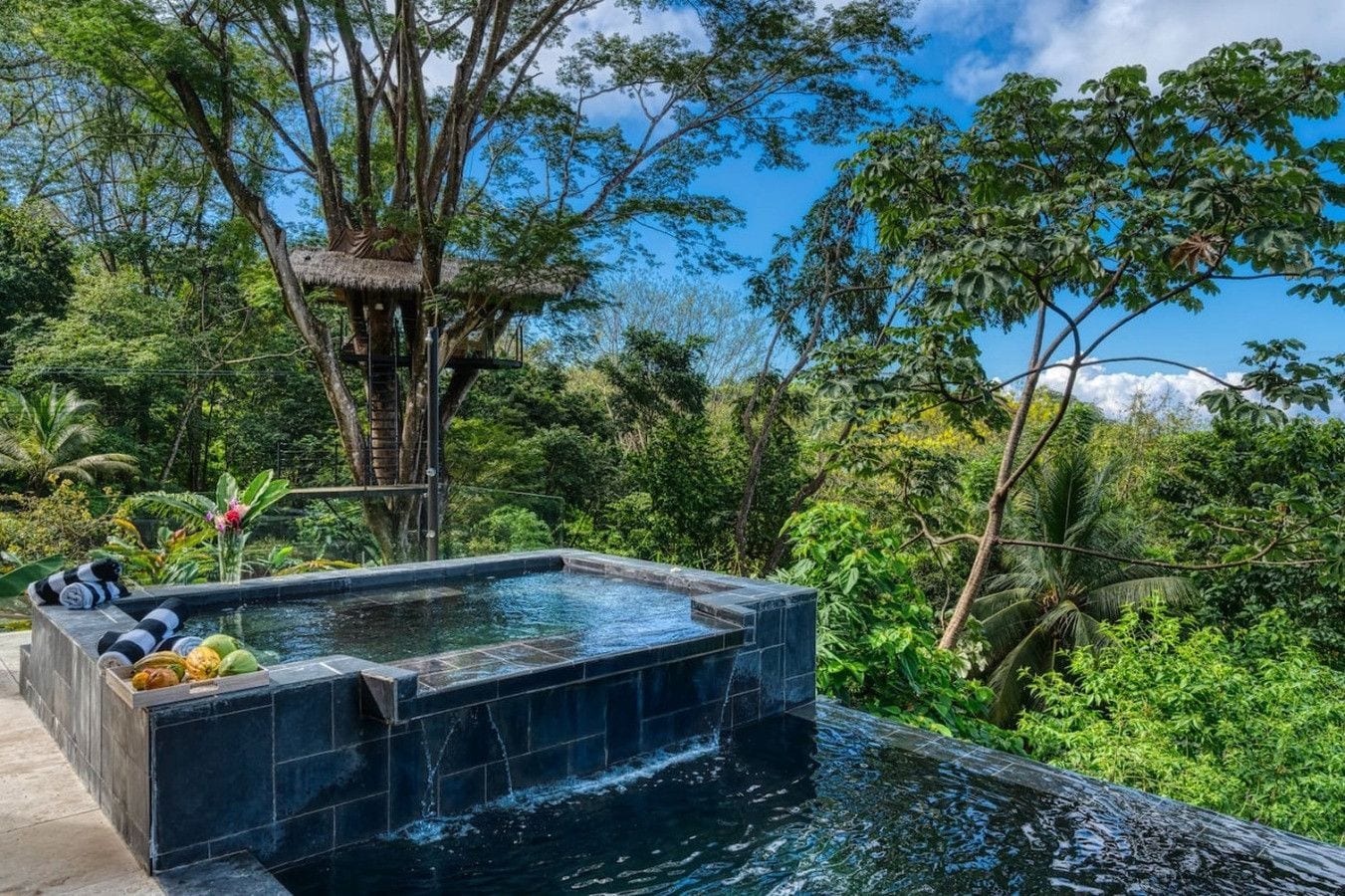 A hot tub with a waterfall feature, surrounded by lush greenery and a thatched-roof structure.
