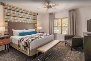 Quarto de hotel com uma cama grande, um banco, uma cadeira, uma escrivaninha, um ventilador de teto e uma janela com cortinas.