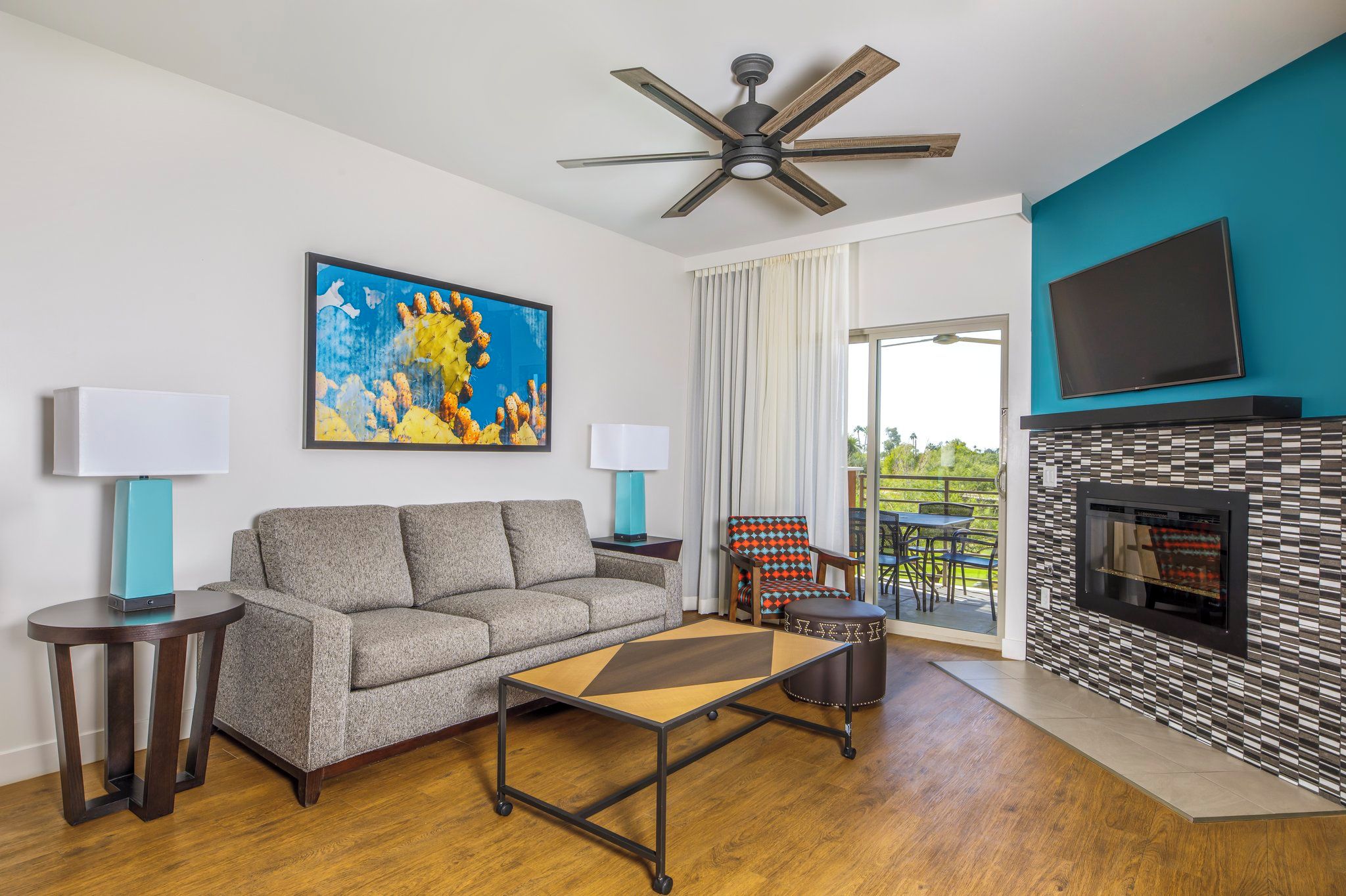 A modern living room with a sofa, coffee table, and a fireplace. There is a ceiling fan, a TV mounted on the wall, and a painting on the wall.