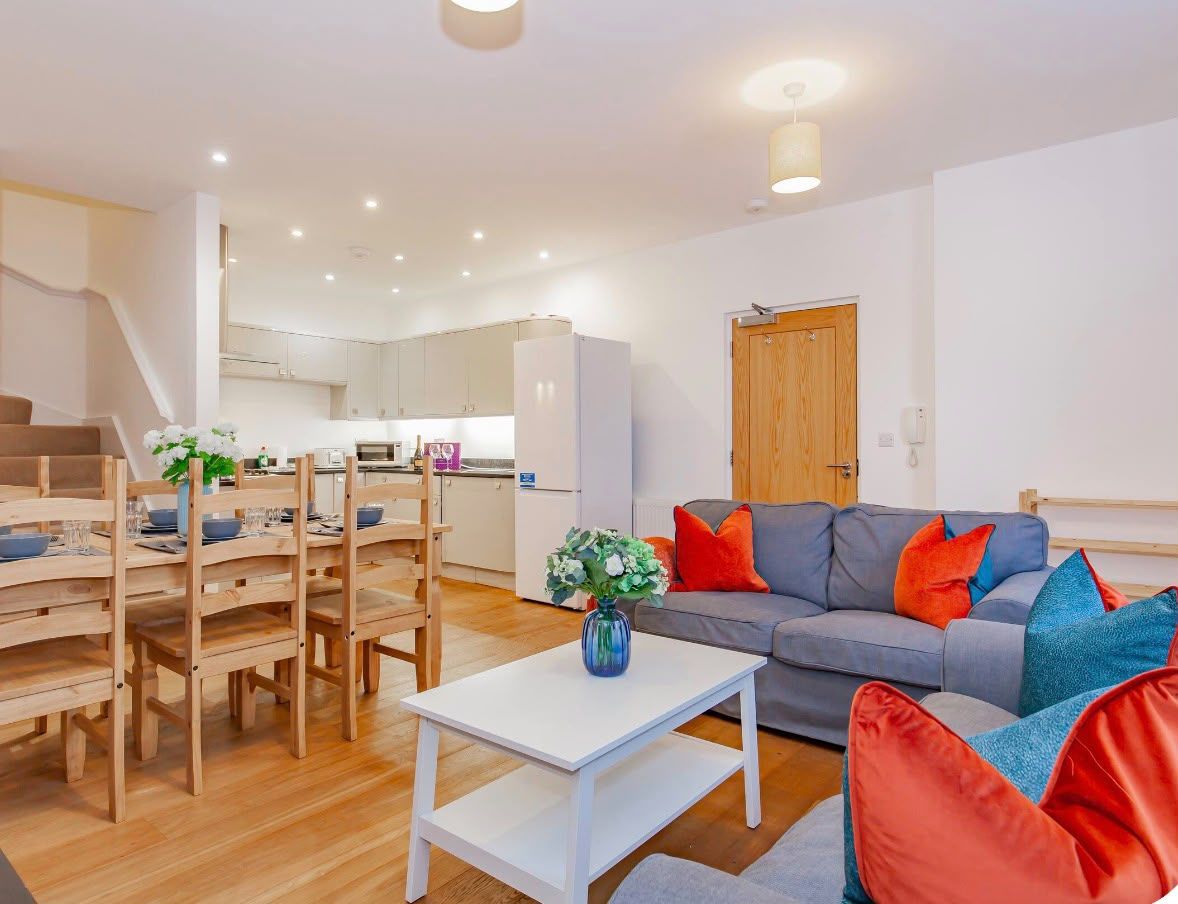 A modern living room with a grey sofa, red and blue cushions, a white coffee table, and a wooden dining table with chairs.