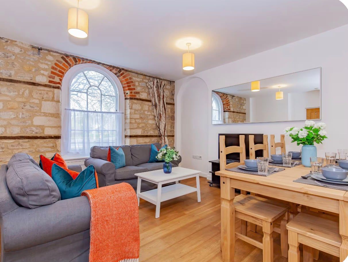 A modern living room with a stone wall, a grey sofa, a wooden dining table set for four, and a large window.