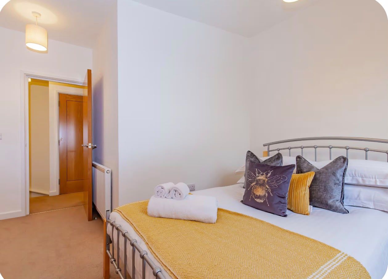 A bedroom with a bed, pillows, a rolled towel, a radiator, and a door leading to another room.