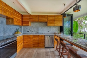 A modern kitchen with wooden cabinets, a granite countertop, and a built-in dishwasher. - Bali Villa Jungle Paradise by Pura Villas (Bahía Ballena)