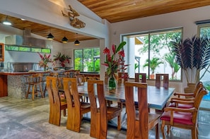 A spacious dining area with wooden chairs and tables, a kitchen with a stainless steel range hood, and large windows offering a view of greenery. - Miralago Villa by Pura Villas (Bahía Ballena)