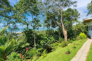 A lush garden with a pathway leading to a building, surrounded by dense foliage and a view of the sea. - Miralago Villa by Pura Villas (Bahía Ballena)