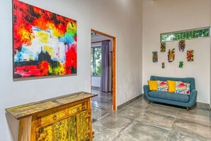 A room with a colorful abstract painting, a wooden cabinet, a blue sofa with patterned cushions, and a window with decorative items. - Miralago Villa by Pura Villas (Bahía Ballena)