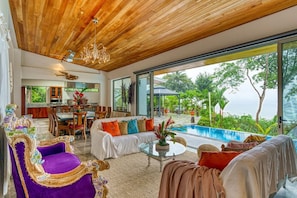 A spacious living area with a pool, wooden ceiling, and large windows overlooking greenery. - Miralago Villa by Pura Villas (Bahía Ballena)