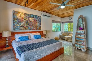 Um quarto espaçoso com uma cama grande, um ventilador de teto, uma pintura abstrata colorida, uma escada de madeira e vista para o verde pelas janelas.