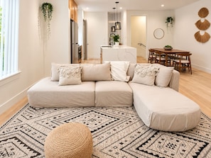 Ein modernes Wohnzimmer mit einer Eckcouch, einem Esstisch mit Stühlen, einer Küchenzeile und einem gemusterten Teppich.
