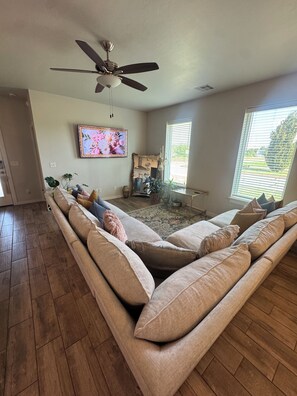 A spacious living room with a large sectional sofa, a ceiling fan, a television, and a window with blinds. - 3-bedroom house in gorgeous Oklahoma City with AC (Oklahoma City)