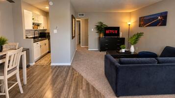A modern living room with a dark sofa, a kitchen area with white cabinets, and a dining table with chairs.