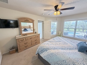 Une chambre à coucher comprenant un lit, une commode, un miroir, un ventilateur de plafond et un téléviseur fixé au mur.