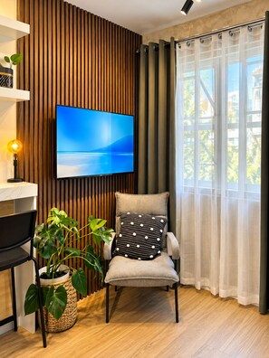 Een moderne woonkamer met een flatscreen televisie aan een houten wand, een comfortabele fauteuil met een gestreept kussen, een bijzettafeltje met een lamp, en een potplant.